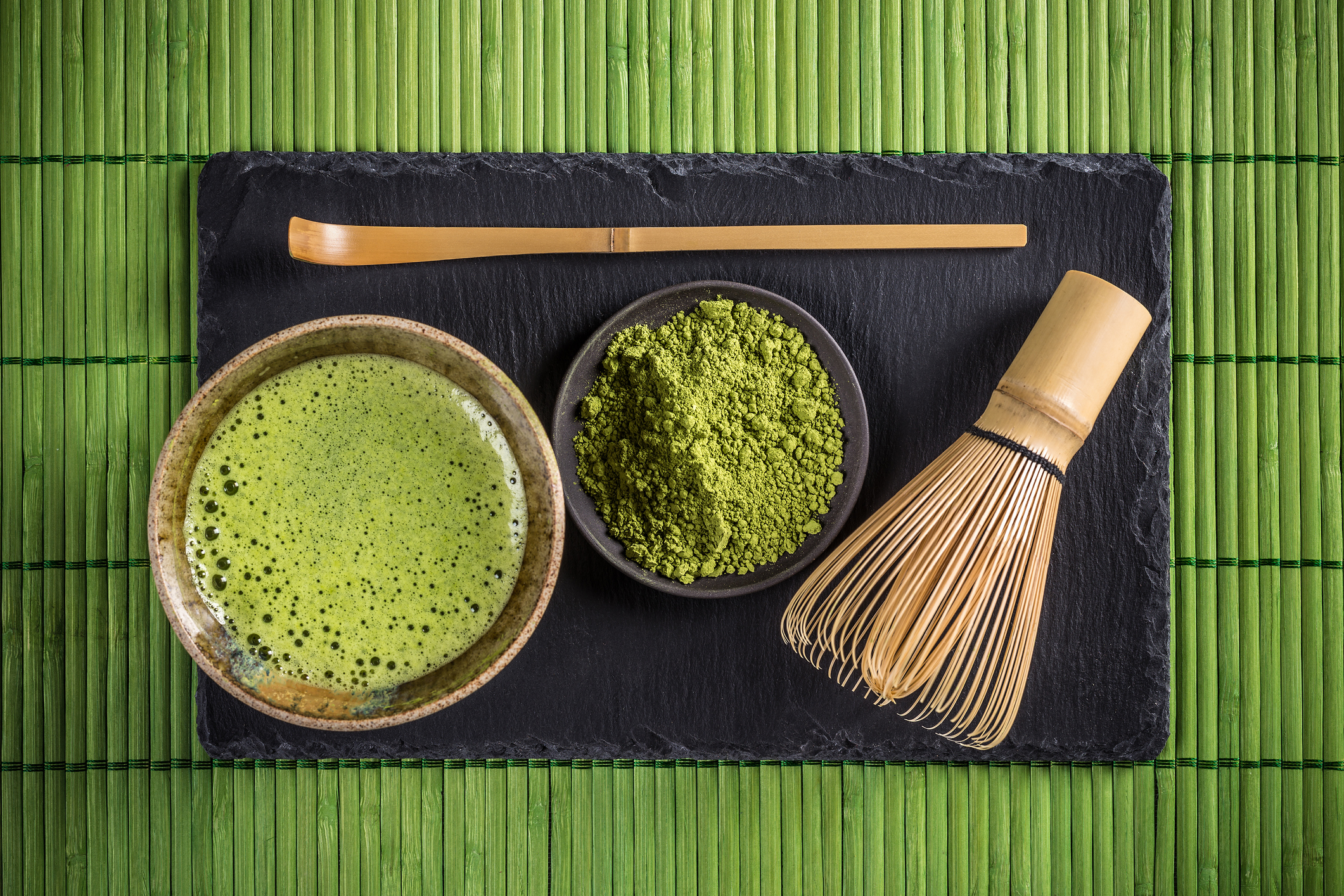 Freshly brewed matcha inside a chawan (tea bowl), next to a plate with matcha green tea powder, a chashaku (tea scoop), and chasen (tea whisk).
